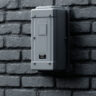A high-quality, professional image showing a secured, tamper-resistant Modern Bait Station placed strategically along the perimeter wall of a commercial building (warehouse or hotel). The station should be discreet but clearly visible, illustrating the safe and controlled use of rodenticides for Pest control in Ghana. The image should convey security and professionalism.